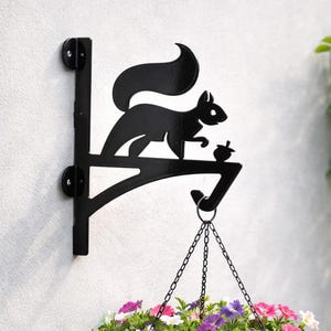 Könnte beinhalten: Schwarzer Metall-Eichhörnchen-Hängekorbhalterung. Die Halterung zeigt eine Silhouette eines Eichhörnchens, das eine Eichel hält. Eine Kette ist an der Halterung befestigt und hält einen Hängekorb mit bunten Blumen. Die Halterung ist an einer weißen Wand montiert.