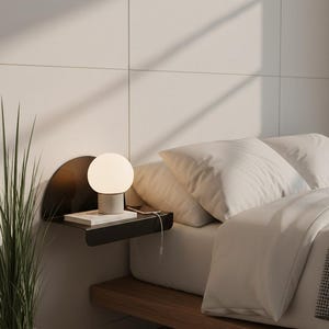 Puede incluir: Una escena de dormitorio moderno con un diseño minimalista. Una lámpara de globo blanca y redonda se encuentra sobre una estantería negra con un libro y un teléfono. La estantería está fijada a una pared blanca, junto a una cama con ropa de cama blanca y un marco de madera.