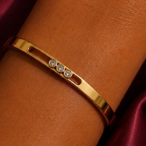 Pode incluir: Uma pulseira rígida de tom dourado com uma ranhura horizontal recortada na superfície frontal. Dentro desta ranhura alongada, três pequenas pedras redondas e transparentes estão engastadas em fila, criando um detalhe decorativo.