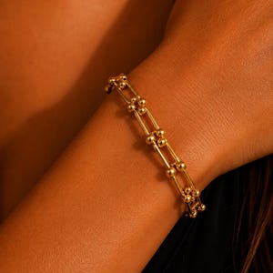 Könnte beinhalten: Ein goldfarbenes Armband mit einer Reihe länglicher, rechteckiger Glieder, die durch kleine, kugelförmige Perlen verbunden sind. Das Design besteht aus zwei parallelen Reihen dieser Glieder und erzeugt eine strukturierte, kettenartige Optik.