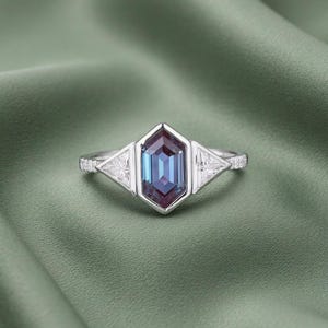 Könnte beinhalten: Ein silberner Ring mit einem sechseckigen, farbwechselnden Edelstein in Blau- und Lilatönen. Zwei dreieckige, weiße Edelsteine flankieren den Mittelstein. Der Ring ist mit kleinen, klaren Akzentsteinen besetzt, vor einem grünen Hintergrund.