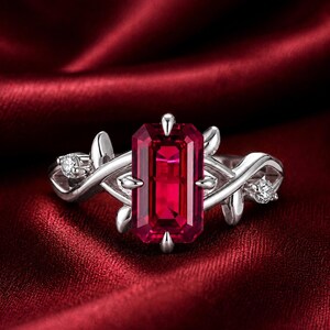 May include: A silver ring featuring a large, rectangular, deep red gemstone as the centerpiece. The ring has a floral vine design with small accent stones on either side of the main stone. The ring is set against a rich, red fabric background.