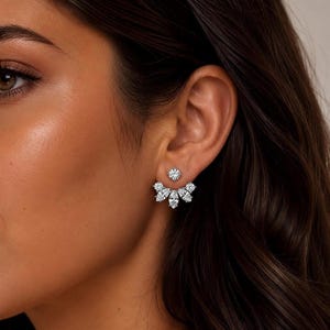 Op de afbeelding: Close-up van een oor van een vrouw versierd met twee witte diamanten oorbellen. De voorste oorbel is een cluster van marquise-geslepen en ronde diamanten. De achterste oorbel is een enkele ronde diamant. De oorbellen zijn van zilver metaal.