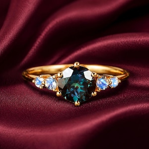 May include: A gold ring with a large, dark teal gemstone at the center, flanked by smaller, light blue stones. The ring sits on a deep red satin surface, creating a luxurious and elegant aesthetic. The ring's band is a simple, polished gold.