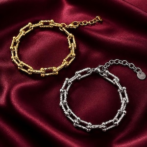 Op de afbeelding: Twee schakelarmbanden, één in goudkleur en één in zilverkleur, met een getextureerd oppervlak op elke rechthoekige schakel en kleine bolvormige accenten bij de verbindingen. Elke armband is voorzien van een verstelbare kettingverlenging.