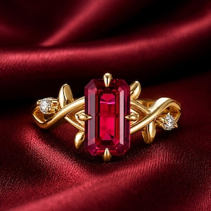 Puede incluir: Un anillo de oro con una gran gema roja rectangular como pieza central. El anillo presenta un diseño en forma de vid con pequeños diamantes a ambos lados de la piedra principal. El anillo está sobre un fondo rojo oscuro.