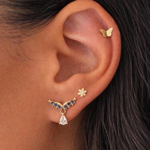 Può includere: Primo piano di un orecchio adornato da orecchini d'oro. L'orecchio presenta un orecchino a farfalla, un piccolo orecchino a fiore e un orecchino d'oro con gemme blu e una gemma a forma di goccia. Gli orecchini sono sulla pelle.