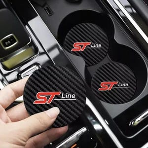 Könnte beinhalten: Schwarze Carbonfaser-Getränkeuntersetzer mit rotem "ST Line"-Logo. Die Untersetzer sind rund und passen in die Getränkehalter eines Autos.