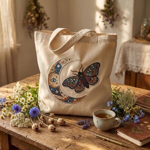 Può includere: Una borsa tote in tela beige con un design ricamato a forma di luna crescente e falena. La luna è blu e oro con un occhio dettagliato. La falena è marrone, blu e oro. La borsa ha due manici ed è su un tavolo di legno.