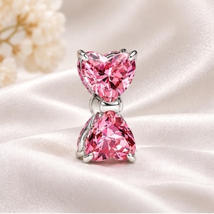 May include: A silver-toned charm featuring two faceted pink heart-shaped stones stacked vertically. The stones are held in place by silver prongs, with a small silver connector piece between the two hearts, creating a bow-like appearance.