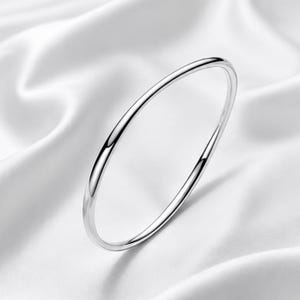 Pode incluir: Uma pulseira fina de tom prateado polido com uma superfície lisa e arredondada. A peça de joalharia circular apresenta um acabamento metálico reflexivo que capta a luz, criando uma estética limpa e simples para este acessório de pulso.