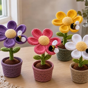 May include: Four crocheted potted flowers in purple, pink, yellow, and white, each featuring a central yellow disc, green stem with two leaves, and a small black and yellow bumblebee attached to a petal, all sitting in matching crocheted pots.