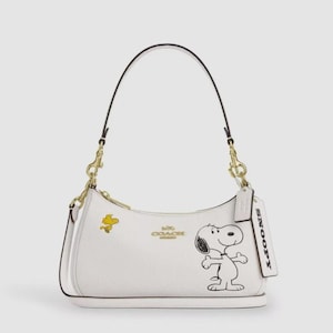 May include: A white leather shoulder bag featuring a graphic of Snoopy and Woodstock. The bag has gold-tone hardware, a zipper closure, and a leather hangtag with the text SNOOPY embossed on it. The Coach logo is printed in gold on the front.
