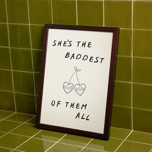 May include: A framed print with the text "SHE'S THE BADDEST OF THEM ALL" in black. The print features a drawing of two cherries with flames inside the hearts. The frame is dark brown.