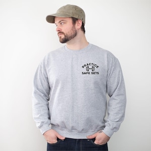 Könnte beinhalten: Hellgraues Sweatshirt mit dem Text "Practice Safe Sets" und einer schwarzen Hantel-Grafik. Das Sweatshirt hat einen Rundhalsausschnitt. Die Person auf dem Bild trägt eine grüne Kappe und blaue Jeans.