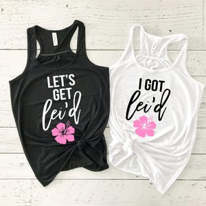 May include: Two tank tops, one black and one white, with the text "LET'S GET lei'd" and "I GOT lei'd" in white and black script, respectively. Each shirt features a pink hibiscus flower graphic. The tank tops are knotted at the bottom.