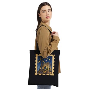 Puede incluir: Un bolso tote negro con un gráfico rectangular diseñado como un sello postal. La imagen muestra la Torre Eiffel y el Arco del Triunfo en París de noche, iluminados por guirnaldas de luces sobre un cielo de color azul oscuro.