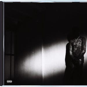 May include: A black and white album cover featuring a person standing in a dimly lit room. The person is wearing a long-sleeved shirt with text. The album cover is in a jewel case.