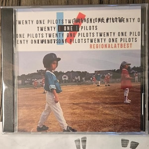 May include: CD case for the album "Regional at Best" by Twenty One Pilots. The cover features a child in a baseball uniform walking on a baseball field. The band's name and album title are printed in white.