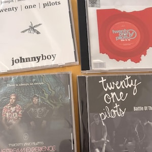 Puede incluir: Cuatro CDs de la banda Twenty One Pilots. El CD superior izquierdo tiene el texto "tyler joseph | twenty | one | pilots" y "johnnyboy". El CD superior derecho tiene un disco rojo con forma de Ohio. Los CDs inferiores tienen fotos y texto de la banda.