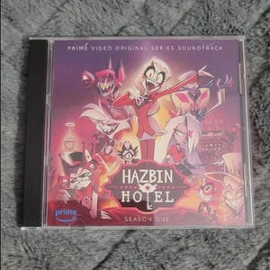 May include: A Hazbin Hotel Season One soundtrack CD in a clear jewel case. The cover features colorful cartoon characters and the text "Hazbin Hotel" and "Season One". The Prime Video logo is visible.
