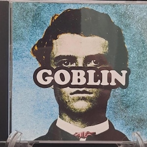 May include: CD case featuring a vintage-style portrait of a man with the word "GOBLIN" obscuring his eyes. The background is a textured blue, and the man is wearing a suit with a bow tie. The CD case is in a clear plastic holder.
