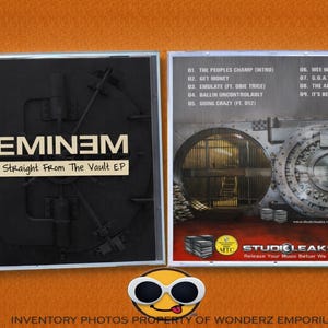 Puede incluir: Caja de CD del álbum "Straight From The Vault EP" de Eminem. La portada es negra con el nombre del artista y el título del álbum en blanco. La parte trasera presenta un diseño de puerta de bóveda y una lista de canciones. Sobre fondo naranja.