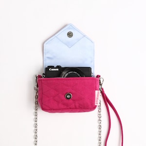 Pode incluir: Uma bolsa tiracolo acolchoada em rosa fúcsia com forro interior azul claro e uma alça de corrente de metal prateado. A bolsa possui um fecho magnético e uma pequena etiqueta branca com o texto mirabell. Uma câmara Canon preta está dentro da bolsa.
