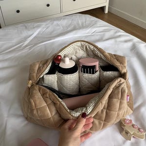 Può includere: Una borsa cosmetica trapuntata beige con cerniera, aperta per rivelare articoli per il trucco. La borsa ha una fodera a quadretti e contiene un rossetto, un porta pennelli trucco e altri prodotti di bellezza. Un compatto rosa e una forcina sono accanto alla borsa.