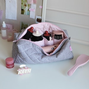 May include: A gingham-patterned cosmetic bag with a pink interior, filled with various beauty tools. A pink hairbrush, lip balm, and a hair clip are also visible. The bag contains brushes, a hair dryer, and other beauty accessories.