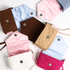 Puede incluir: Una colección de pequeñas bolsas acolchadas con correas de muñeca, cada una con un cierre de broche metálico y una etiqueta 'mirabell'. Las bolsas vienen en varios colores como blanco, marrón, beige, azul y magenta, con forros de satén.