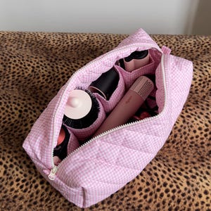 May include: A quilted light pink and white gingham patterned zipped travel case with internal compartments containing various hair styling tools, including a pink cylindrical device with the Dyson brand name printed on the side.