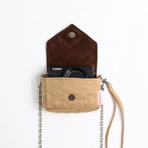 Pode incluir: Uma bolsa tiracolo de lona bege acolchoada com aba interna marrom escura e alça de corrente de metal prateado. A bolsa possui fecho magnético, alça de pulso e uma pequena etiqueta rosa com o texto mirabell. Uma câmera Canon preta.