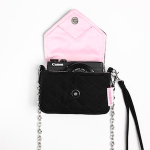 May include: A black quilted fabric pouch with a silver chain strap and a black wrist strap. The interior flap is lined with light pink fabric. A black Canon camera is inside the pouch. A small pink tag with the word mirabell is attached.