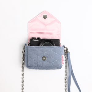 May include: A light blue quilted denim crossbody bag with a pink satin interior lining and a silver metal chain strap. The bag features a magnetic snap closure and a small white tag with the text mirabell. A black Canon camera is inside.