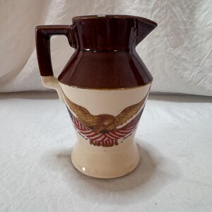 May include: A brown and cream-colored ceramic pitcher with a handle and spout. The pitcher features a design of a golden eagle with outstretched wings, red and white stripes, and blue accents. The words "Spirit of America" are printed on the pitcher.