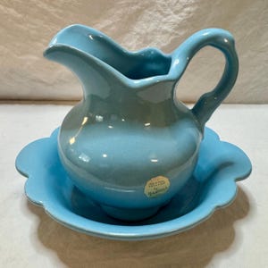 May include: A vintage blue ceramic pitcher and basin set. The pitcher has a curved handle and spout, and sits on a matching scalloped-edge basin. The set is a light blue color with a glossy finish. The label reads "Original Creation by Francoma".