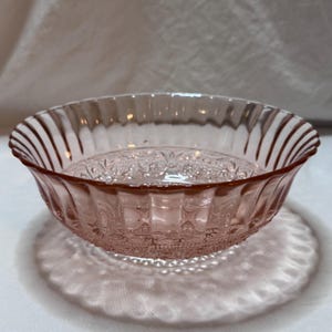 May include: A pink glass bowl with a decorative pattern on the inside and a ribbed exterior. The bowl is round with a scalloped rim and casts a shadow on the surface below.