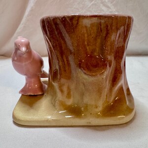 May include: A ceramic toothpick holder shaped like a tree stump with a pink bird perched on the side. The stump is brown with lighter tan accents, and the base is a light tan color.