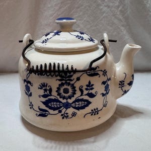 May include: Cream-colored ceramic teapot with a blue floral pattern. The teapot has a lid with a blue knob and a metal handle. The design includes flowers and leaves. The spout is on the right side.