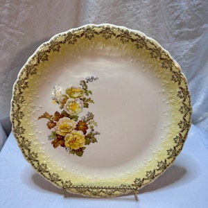May include: A vintage, cream-colored plate with a yellow border and a floral design. The plate features a scalloped edge with gold trim and a bouquet of yellow roses and brown leaves. The plate is displayed on a gold-colored stand.