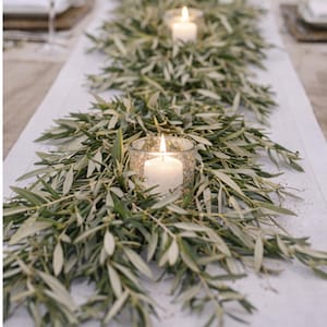 May include: A table runner featuring olive branches and lit candles in glass holders. The green leaves and white candles create a natural, elegant centerpiece. The candles are cylindrical and provide a warm glow.