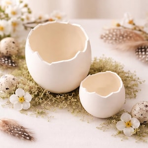 Könnte beinhalten: Zwei weiße, eierschalenförmige Behälter mit gezackten Rändern, einer größer als der andere, liegen auf einem Bett aus grünem Moos. Kleine gesprenkelte Eier, weiße Blumen und Federn umgeben sie und schaffen ein Frühlings- oder Osterthema.