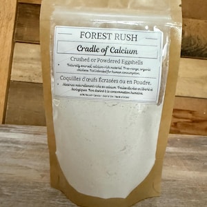 May include: A brown paper bag labelled "FOREST RUSH Cradle of Calcium" containing crushed or powdered eggshells. The label also includes text in French. The product is described as naturally sourced, calcium-rich material from free-range, organic chickens.