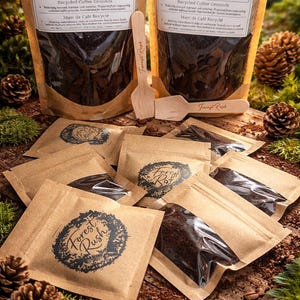 Peut inclure: Deux grands sachets bruns et plusieurs petits paquets bruns de "FOREST RUSH Rootwake Recycled Coffee Grounds" sont présentés. Des cuillères en bois avec le logo "Forest Rush" sont également présentes. Les produits sont disposés sur une surface en bois avec des pommes de pin et de la mousse.