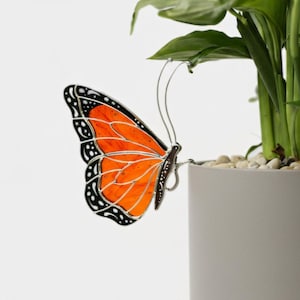 May include: A decorative butterfly with orange and black wings, perched on the edge of a white planter. The butterfly has a silver metal body and is near a green plant. The wings have a stained glass appearance.