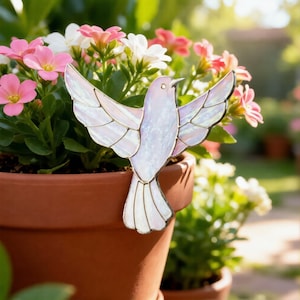 May include: An iridescent glass dove ornament with outstretched wings, placed on a terracotta flower pot. The dove is set against a backdrop of pink and white flowers, creating a tranquil garden scene.