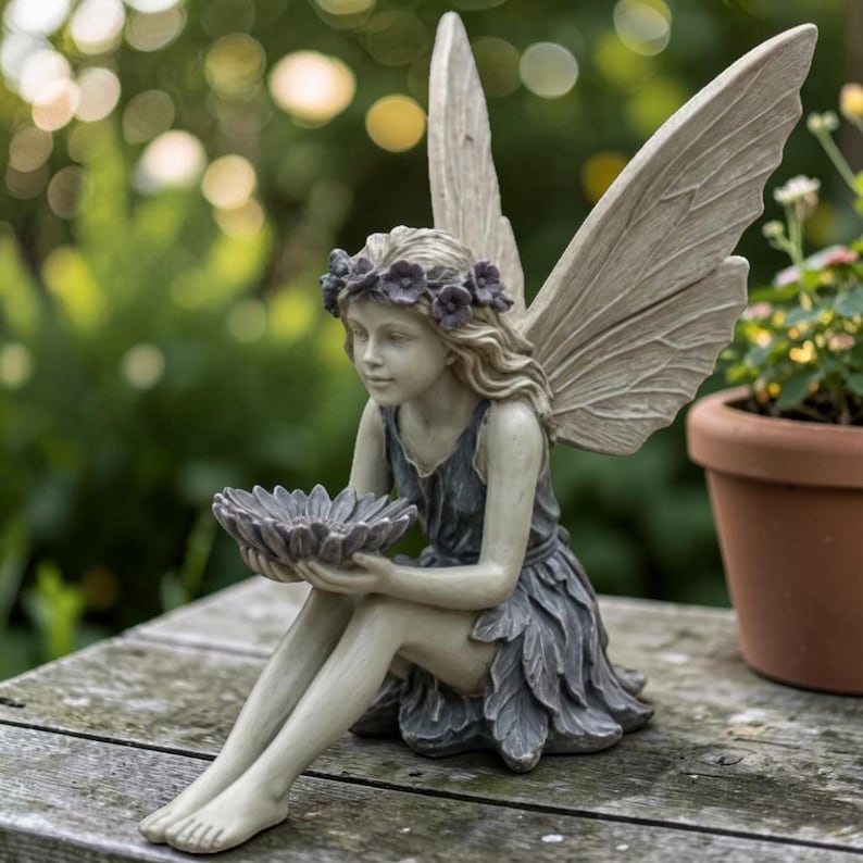 K&ouml;nnte beinhalten: Detaillierte Statue einer sitzenden Fee mit gro&szlig;en, strukturierten Fl&uuml;geln. Die Fee tr&auml;gt ein dunkelblaues Kleid und einen Blumenkranz und h&auml;lt eine graue, bl&uuml;tenf&ouml;rmige Schale. Die Statue besteht aus hellem Material und eignet sich f&uuml;r die Gartendekoration.