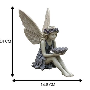 K&ouml;nnte beinhalten: Eine sitzende Feenfigur, etwa 14 cm hoch und 14,8 cm breit, mit gro&szlig;en, detaillierten Fl&uuml;geln. Die Fee tr&auml;gt ein blau-graues Kleid und einen Blumenkranz und h&auml;lt eine bl&uuml;tenf&ouml;rmige Schale. Die Statue besteht aus einem stein&auml;hnlichen Material.