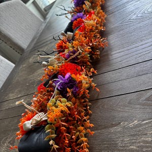 May include: A Halloween-themed table garland featuring artificial orange and brown autumn foliage, purple and red fabric flowers, black branches, and small plastic skeleton figures. The decorative piece is arranged in a long, linear shape.